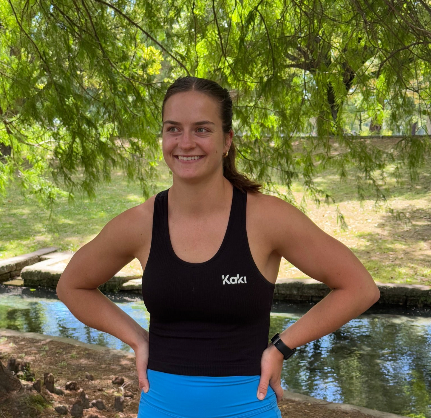A woman standing outdoors wearing a black ribbed racerback tank top with the brand name 'Kaki' visible on the front.
