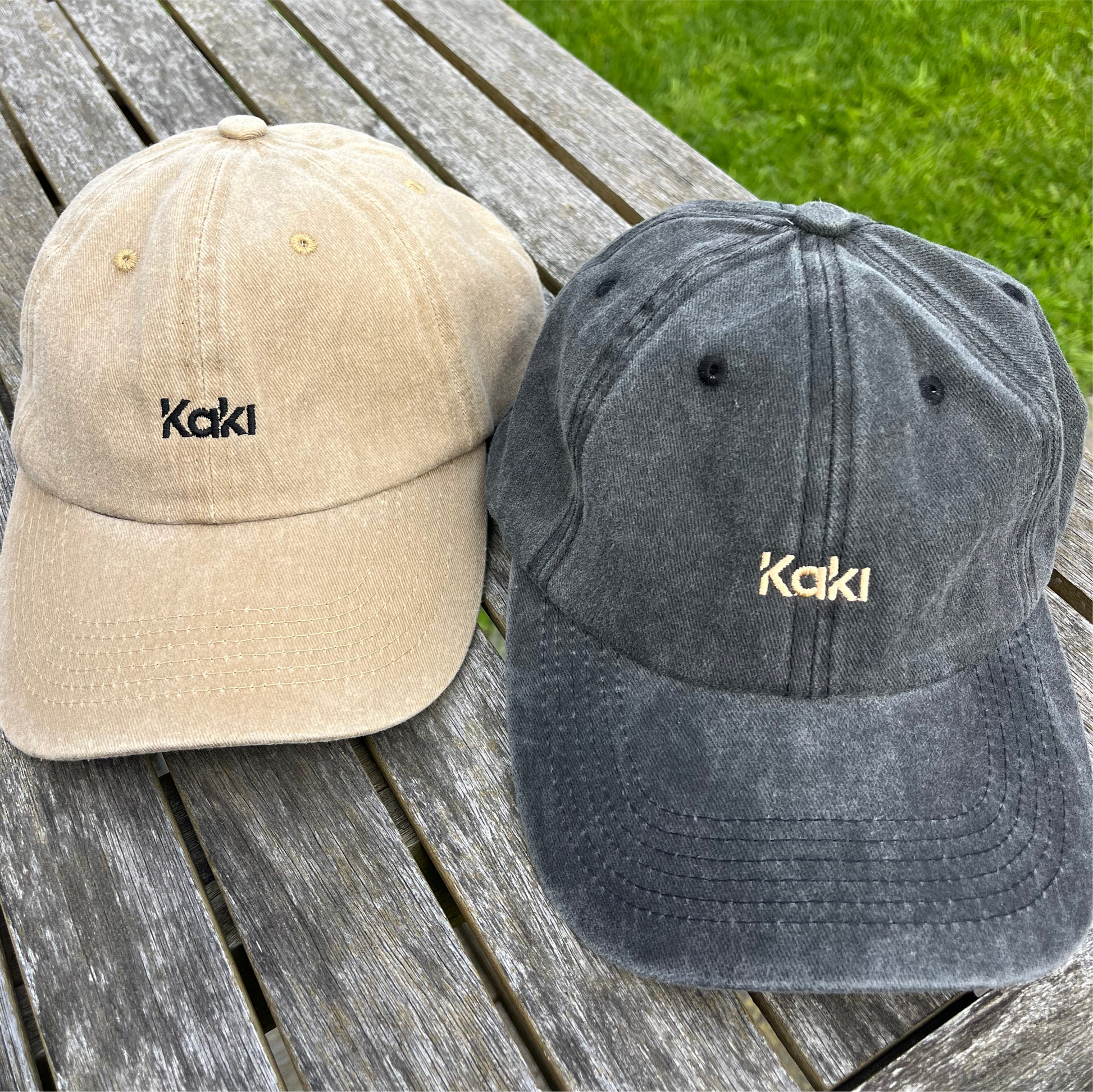 Two baseball caps, one beige and one gray, with 'Kaki' branding on a wooden surface.