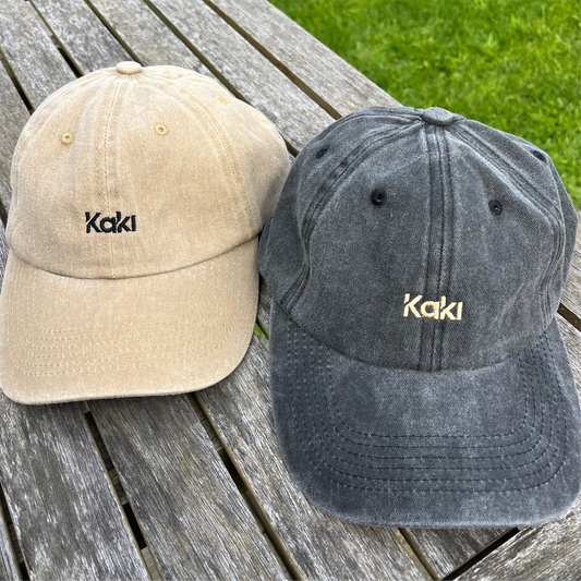 Two baseball caps, one beige and one gray, with 'Kaki' branding on a wooden surface.