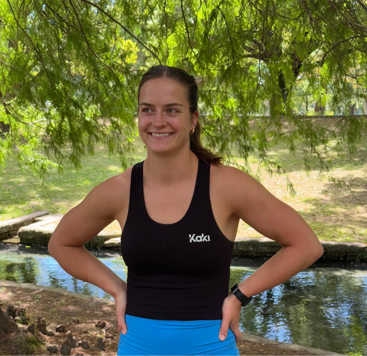 A woman standing outdoors wearing a black ribbed racerback tank top with the brand name 'Kaki' visible on the front.